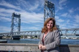 Woman with brown hair wearing gray suit stands in front of Memorial Bridge in Portsmouth.